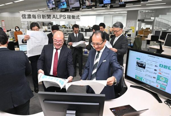  信濃毎日新聞本社の編集フロアで開かれた稼働式で、記念の号外を手にする小坂社長（手前右）、読売新聞東京本社の津田取締役（同左）ら＝2025年12月15日、長野市